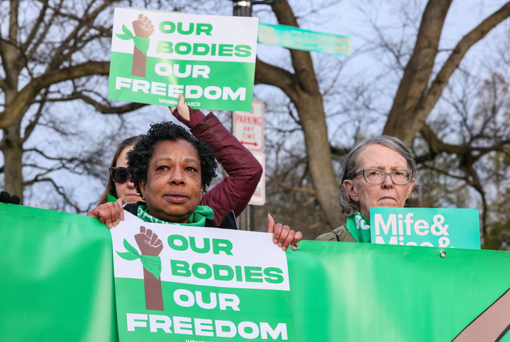 Women at a protest hold green signs that say "OUR BODIES OUR FREEDOM."